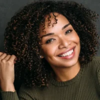 Woman with curly hair wearing an olive green sweater smiles at the camera, holding a strand of her hair with one hand in front of a dark background.