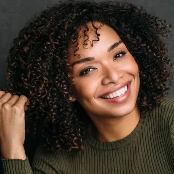 Woman with curly hair wearing an olive green sweater smiles at the camera, holding a strand of her hair with one hand in front of a dark background.