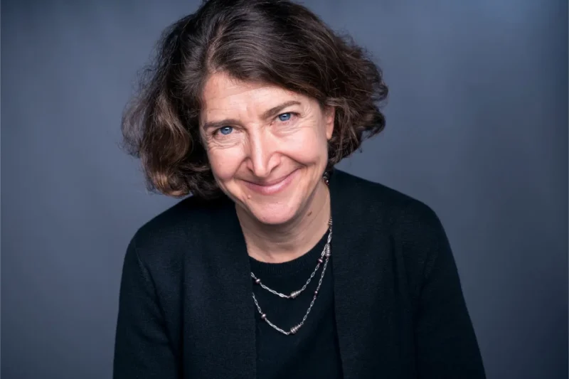Woman with short brown curly hair and a black sweater smiling slightly, wearing a necklace, pictured against a plain gray background.