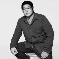 A person wearing glasses and a button-up shirt with a jacket sits on a stool against a plain background, looking at the camera.