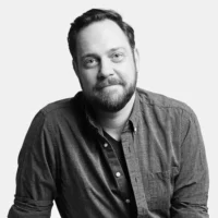 A man with a beard and short hair, wearing a button-up shirt, sits and looks at the camera against a plain background.