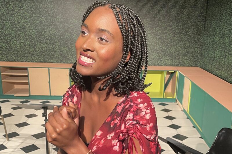 Woman with braided hair wearing a red floral dress stands indoors, clasping her hands and smiling, with green walls and checkered floor in the background.