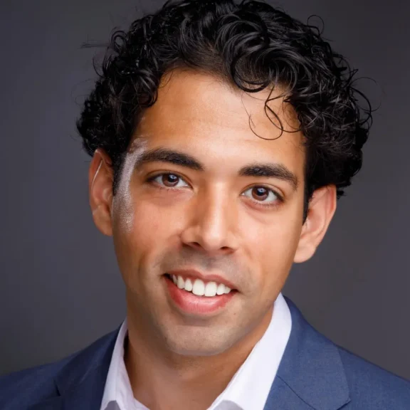 Smiling man with curly dark hair wearing a navy blue blazer and white shirt, photographed against a plain dark background.