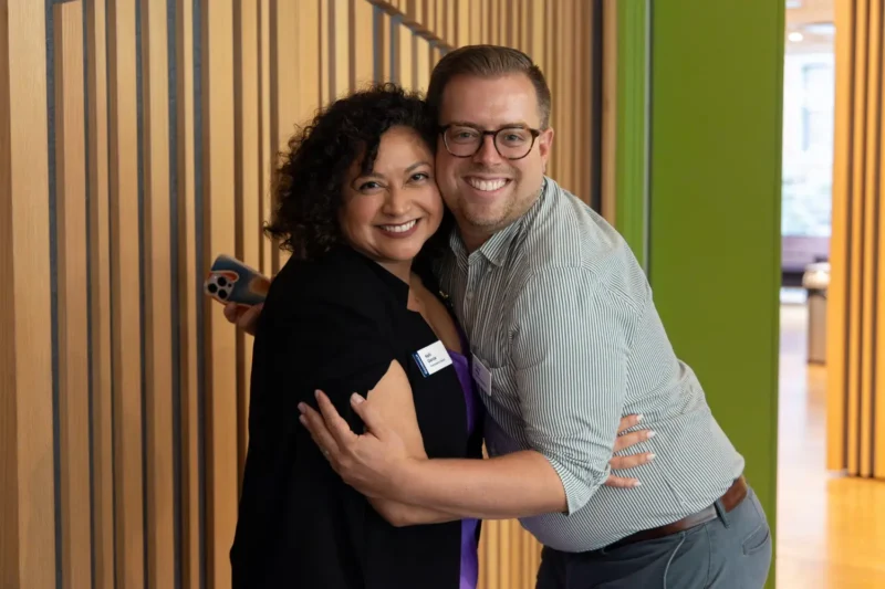 Two people stand indoors, smiling and hugging in front of a wooden paneled wall. One holds a phone, and both wear name tags.