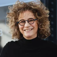 A person with curly hair, round glasses, and a black turtleneck sweater is smiling while standing outdoors.