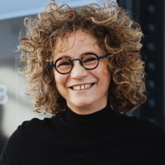 A person with curly hair, round glasses, and a black turtleneck sweater is smiling while standing outdoors.