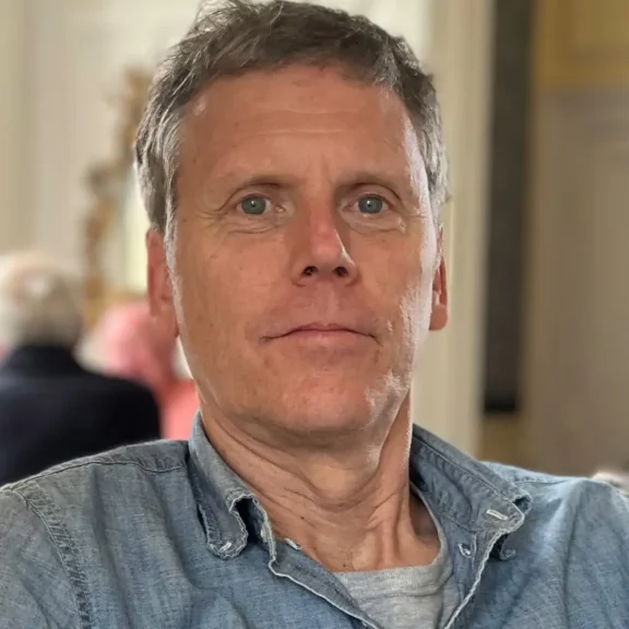 A middle-aged man with short gray hair wears a light blue denim shirt and looks at the camera. There are blurred figures and a light-colored wall in the background.