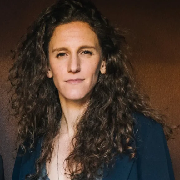 A person with long curly brown hair, wearing a dark jacket, stands in front of a plain brown background, looking directly at the camera with a neutral expression.