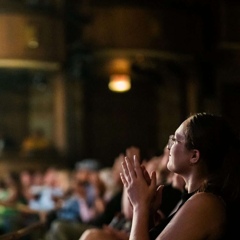A side profile of a woman in a dark theater clapping for a performance.