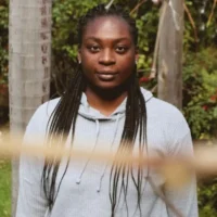 A young Black woman with long braids wearing a gray hooded sweatshirt stands in front of a tree.