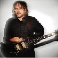 A middle-aged man with glasses and shaggy hair, wearing a dark button-down shirt, holds an electric guitar.