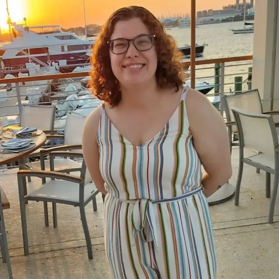 A woman with curly auburn hair and glasses smiling on a patio overlooking a marina at sunset.