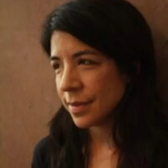 Woman with long dark hair looks slightly to the side, standing against a plain brown wall.