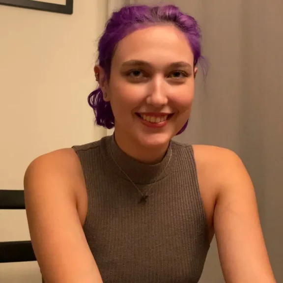 A smiling person with short purple hair wearing a sleeveless grey top.