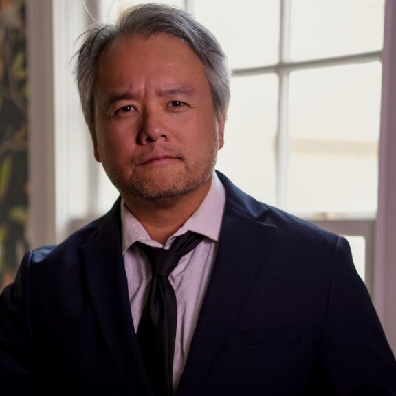 A middle-aged man with gray hair and facial hair, wearing a dark blazer, light shirt, and black tie.