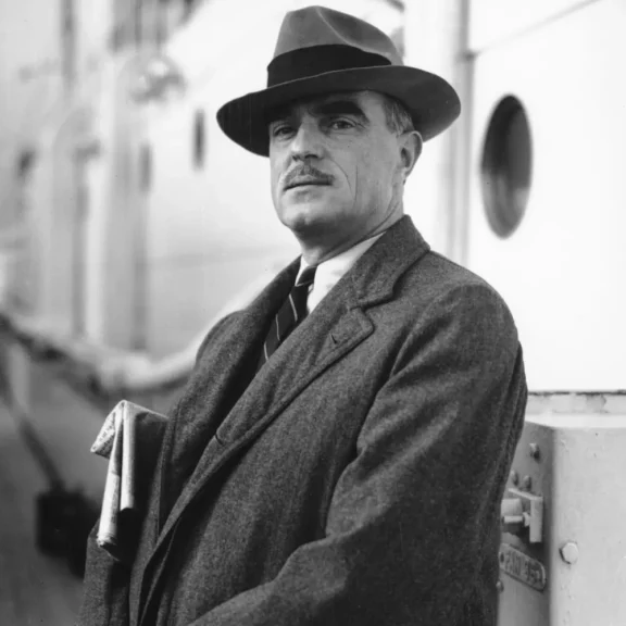A man with a mustache, wearing a fedora and a heavy coat, standing on the deck of a ship.