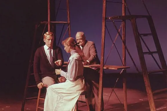 A young man and woman sit on stools on a stage, while an older man sits at a table behind them between two wooden ladders.